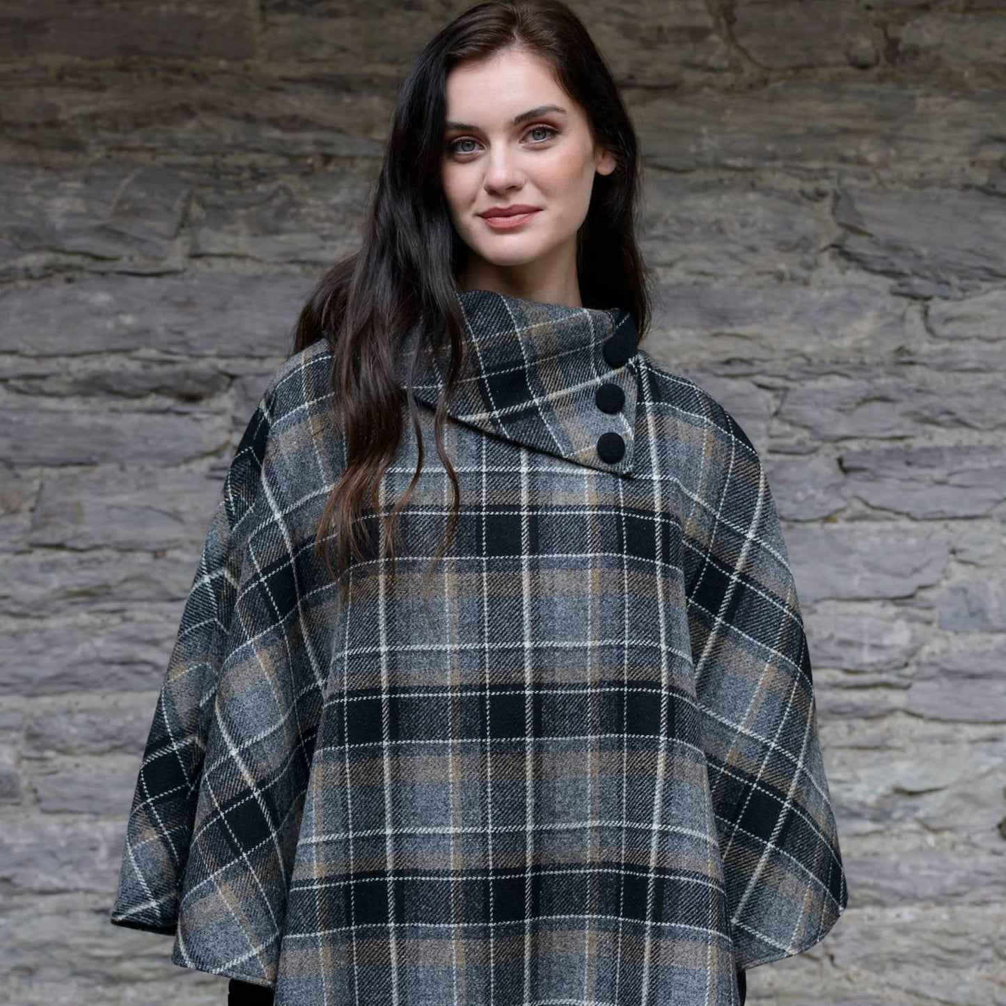 A woman standing against a stone wall background, wearing a gray, black, and beige checkered wool poncho with a collar and buttons.