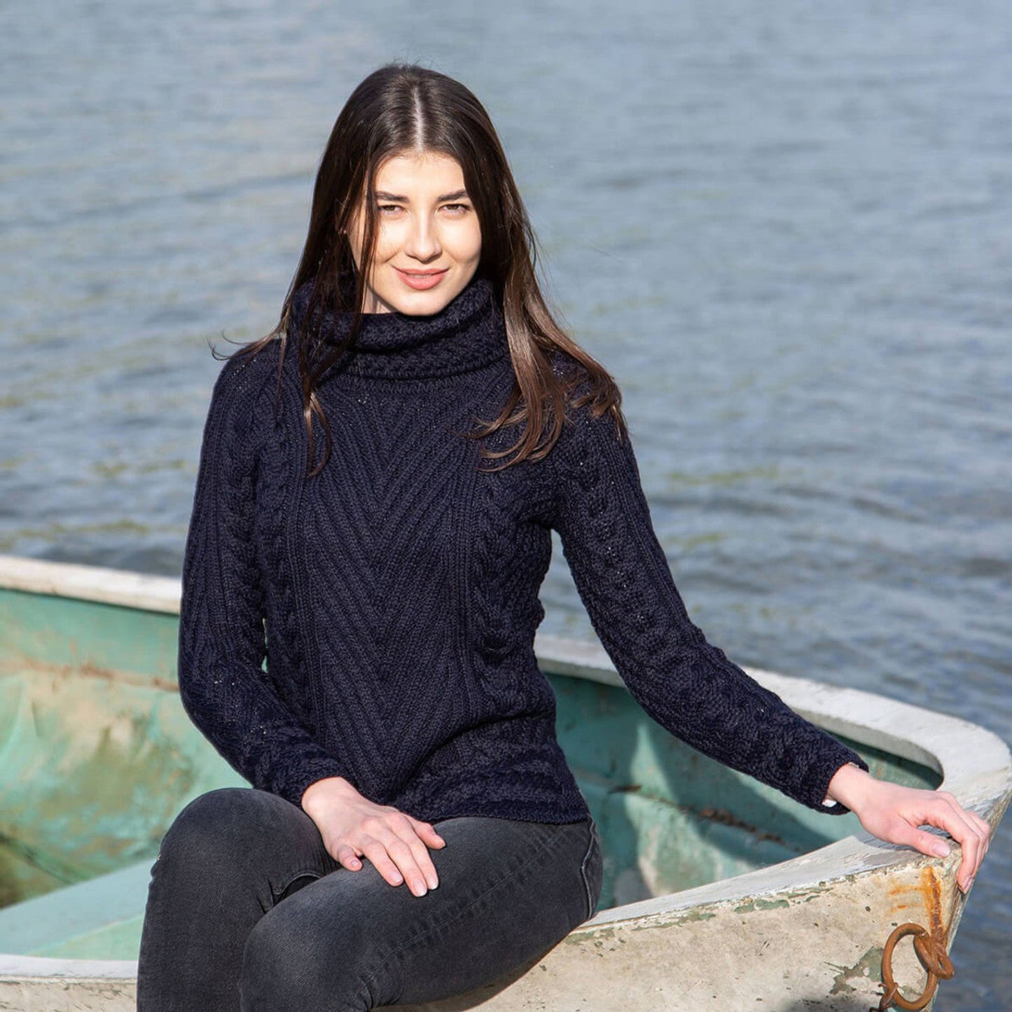 A woman wearing a beige turtleneck ribbed cable knit sweater in an outdoor setting with trees in the background.