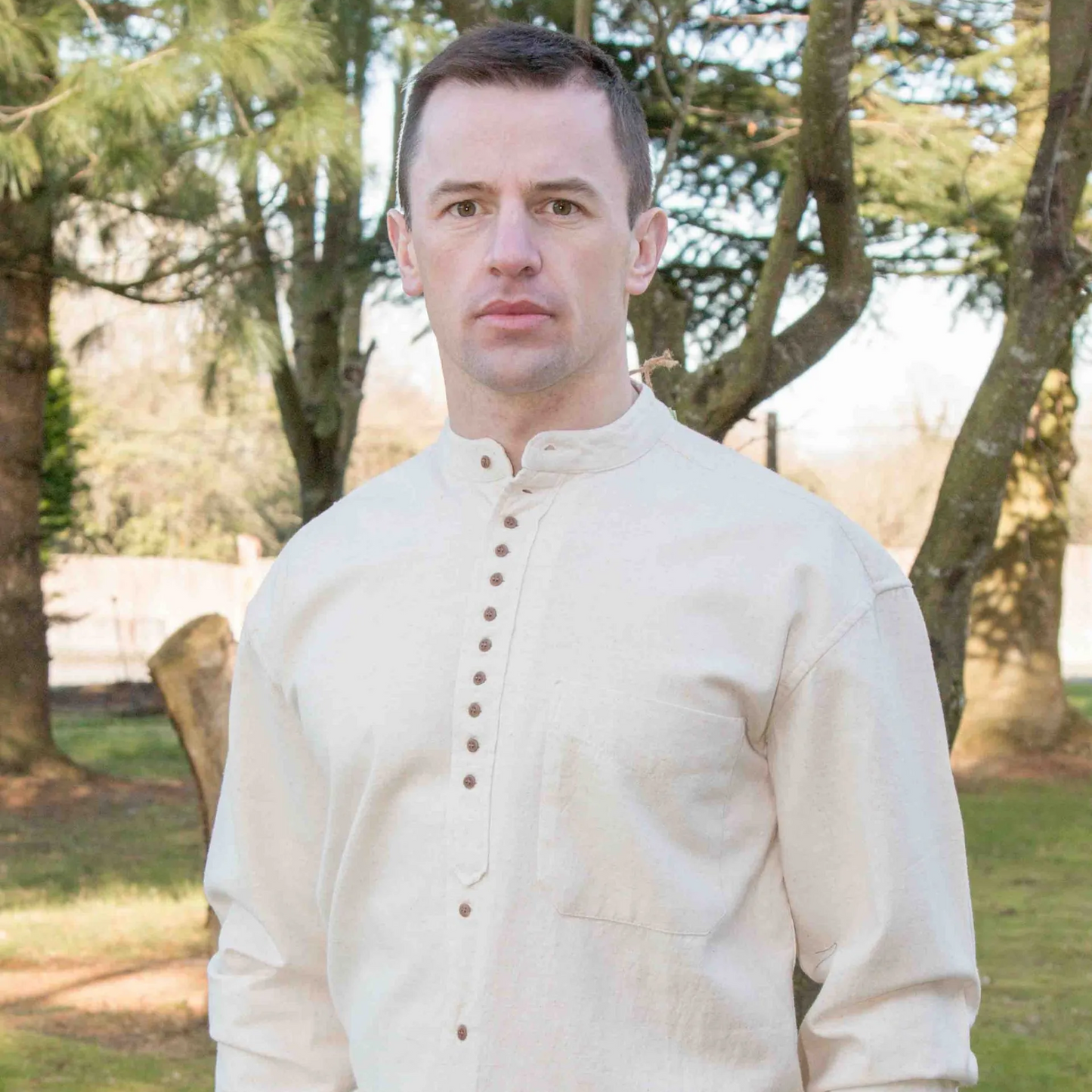 A man standing outdoors wearing a long-sleeved, button-up, beige-colored shirt with a band collar.