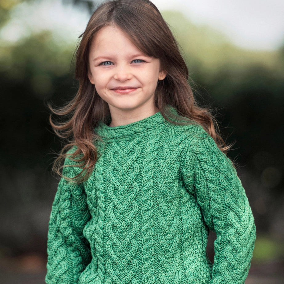 A young girl with long brown hair wearing a green cable-knit sweater and blue jeans, standing outdoors with a blurred natural background.