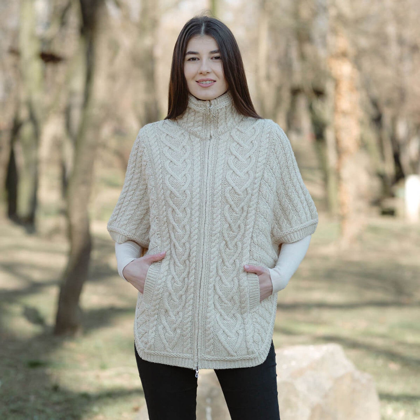 A woman standing outdoors wearing a beige funnel neck poncho jacket with a zipper and side pockets.
