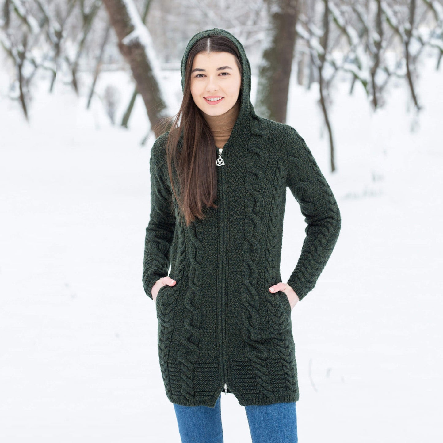 A woman standing outdoors in a snowy setting, wearing a green cable knit hooded zip coatigan with ribbed side pockets.