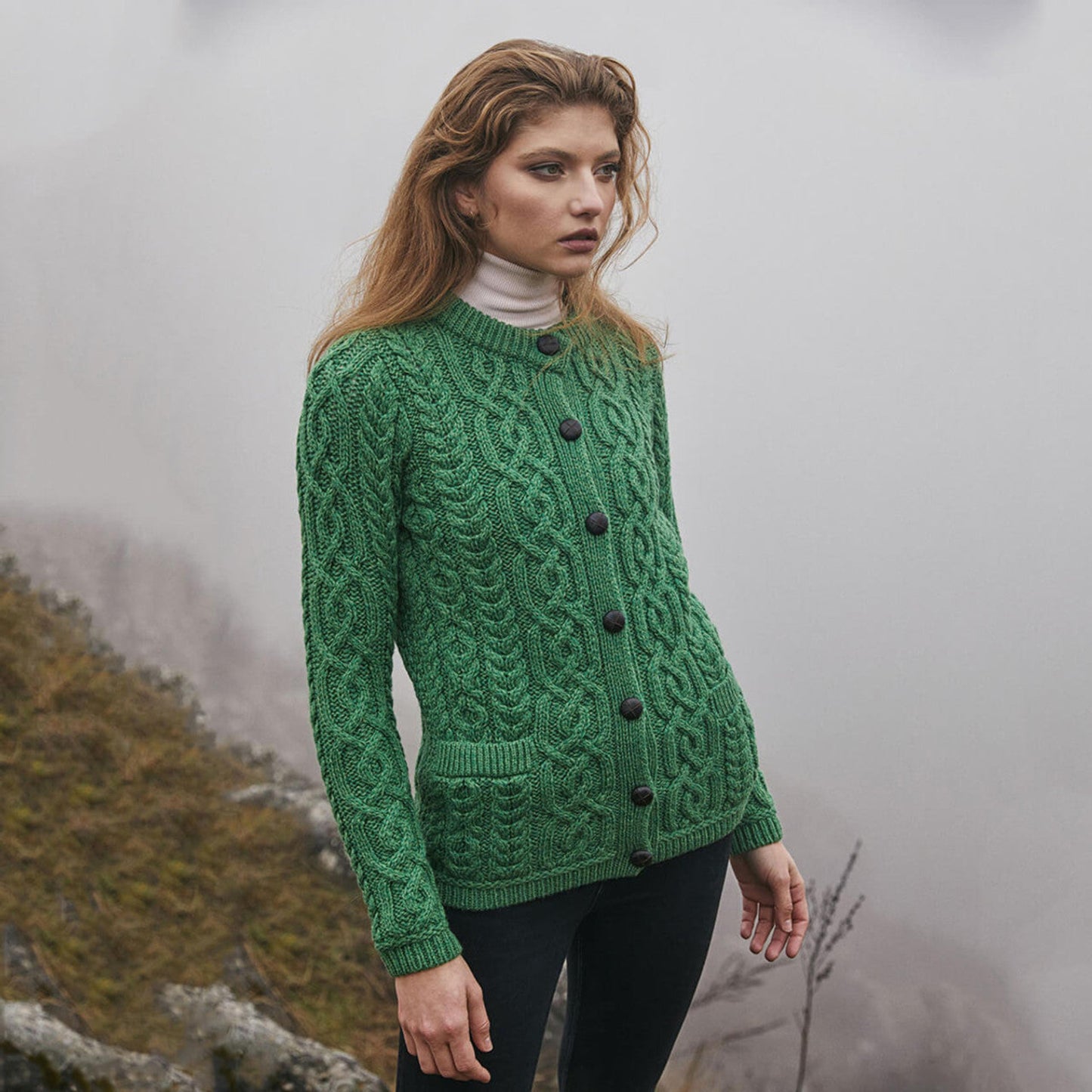 A woman wearing a green traditional Aran cardigan with brown buttons and ribbed cuffs, standing outdoors.
