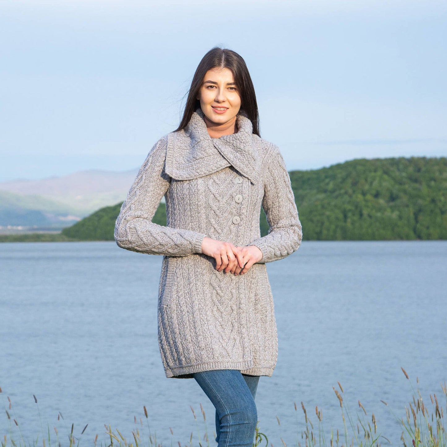 A woman standing outdoors wearing a green, chunky knit cable coat with an asymmetric collar and side fastening.