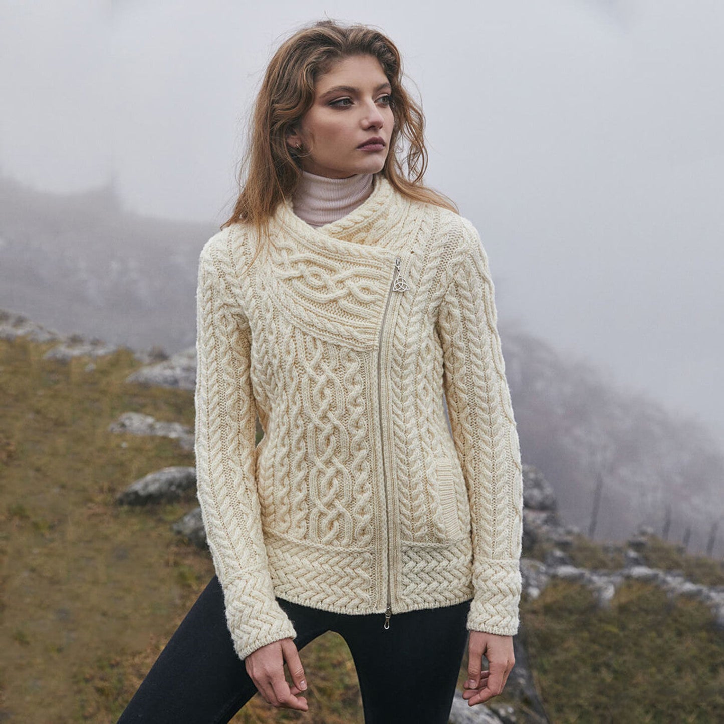 A woman standing outdoors wearing a cream cable knit side zip cardigan with a V-neckline over a white top, paired with black pants.