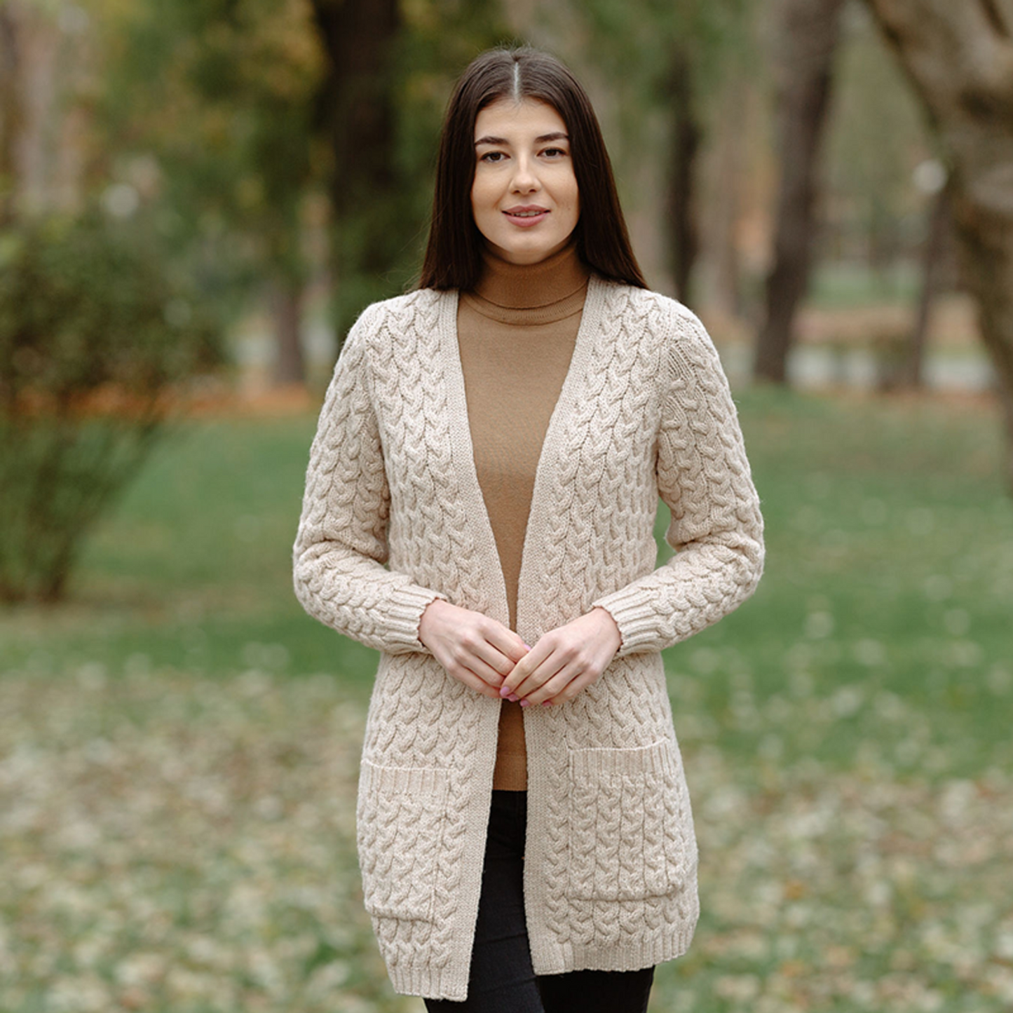A woman standing outdoors wearing a beige Irish cardigan with Aran cable patterns over a brown turtleneck.