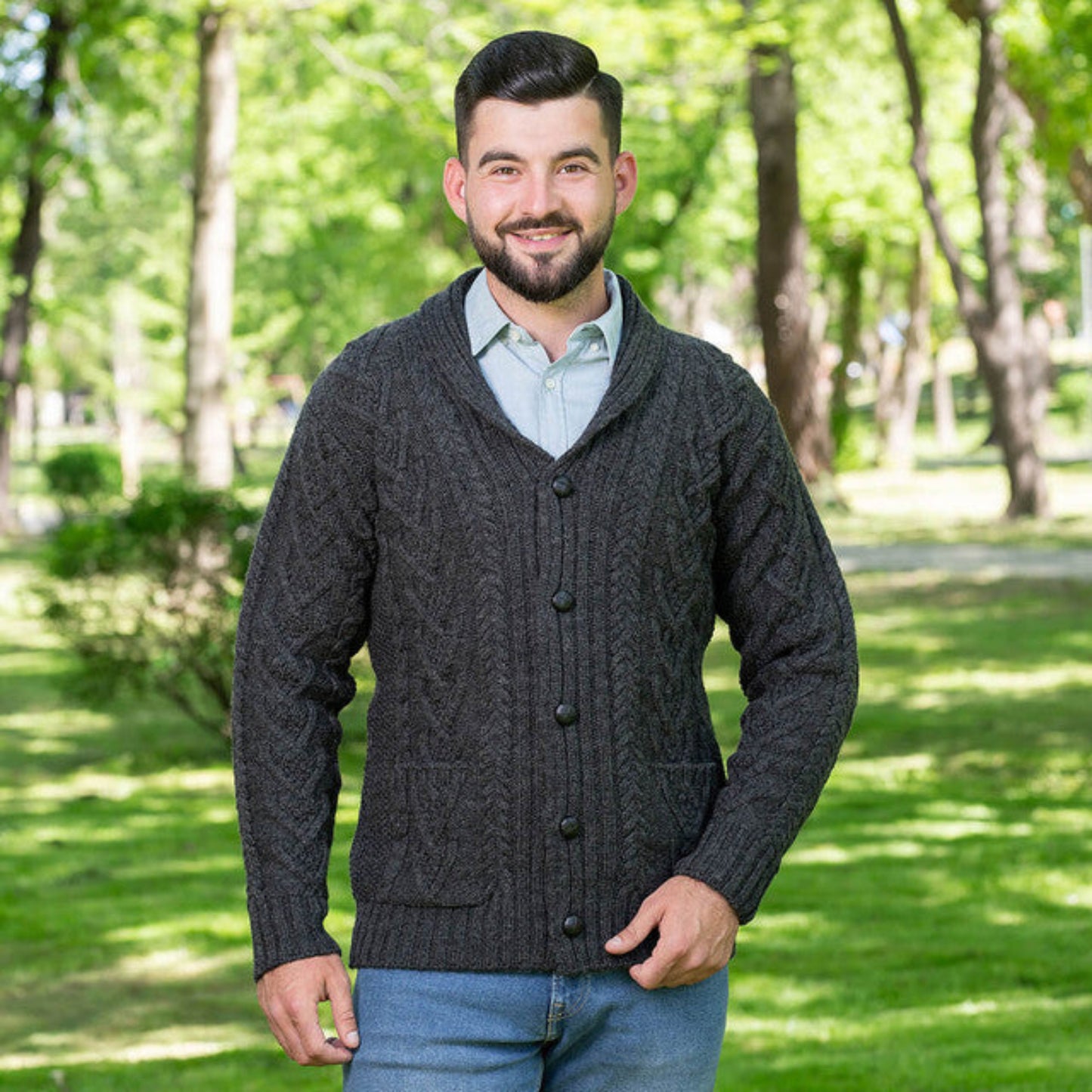 A man wearing a gray shawl-collar aran cardigan with brown buttons, paired with a blue shirt and jeans, standing outdoors.