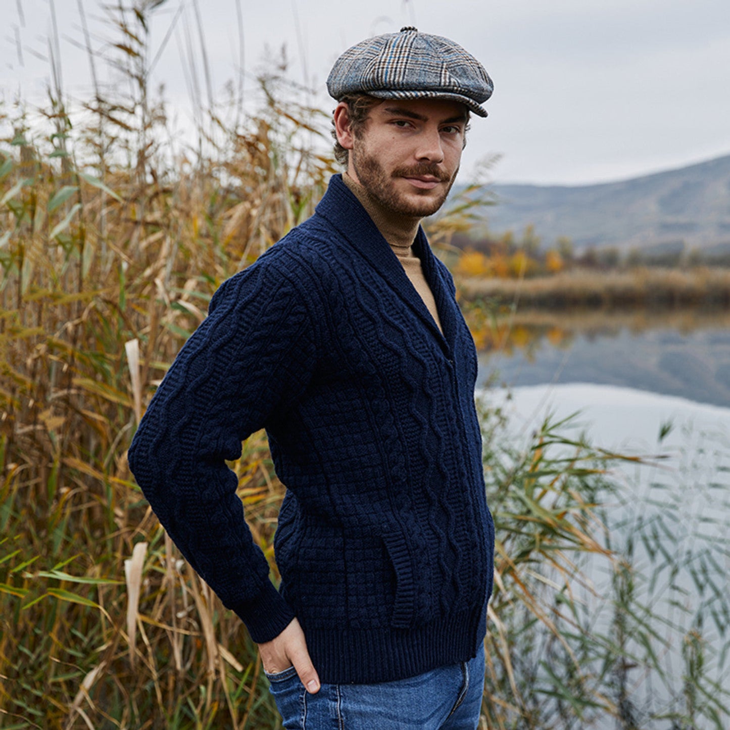 A man standing by a lake wearing a navy blue zip knit cardigan and a checkered flat cap.