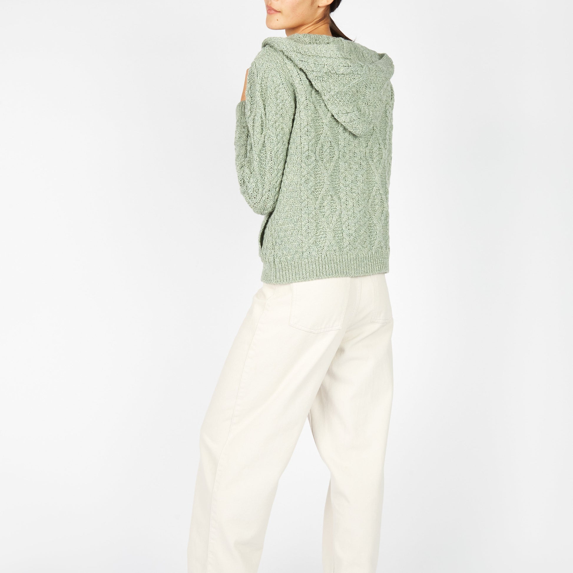 Person wearing a green cable knit sweater and white pants on a white background