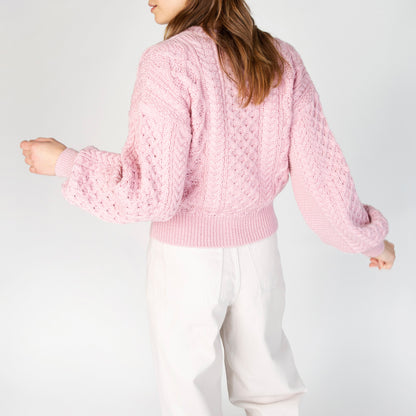 Person wearing a pink cable knit sweater and white pants on a light gray background