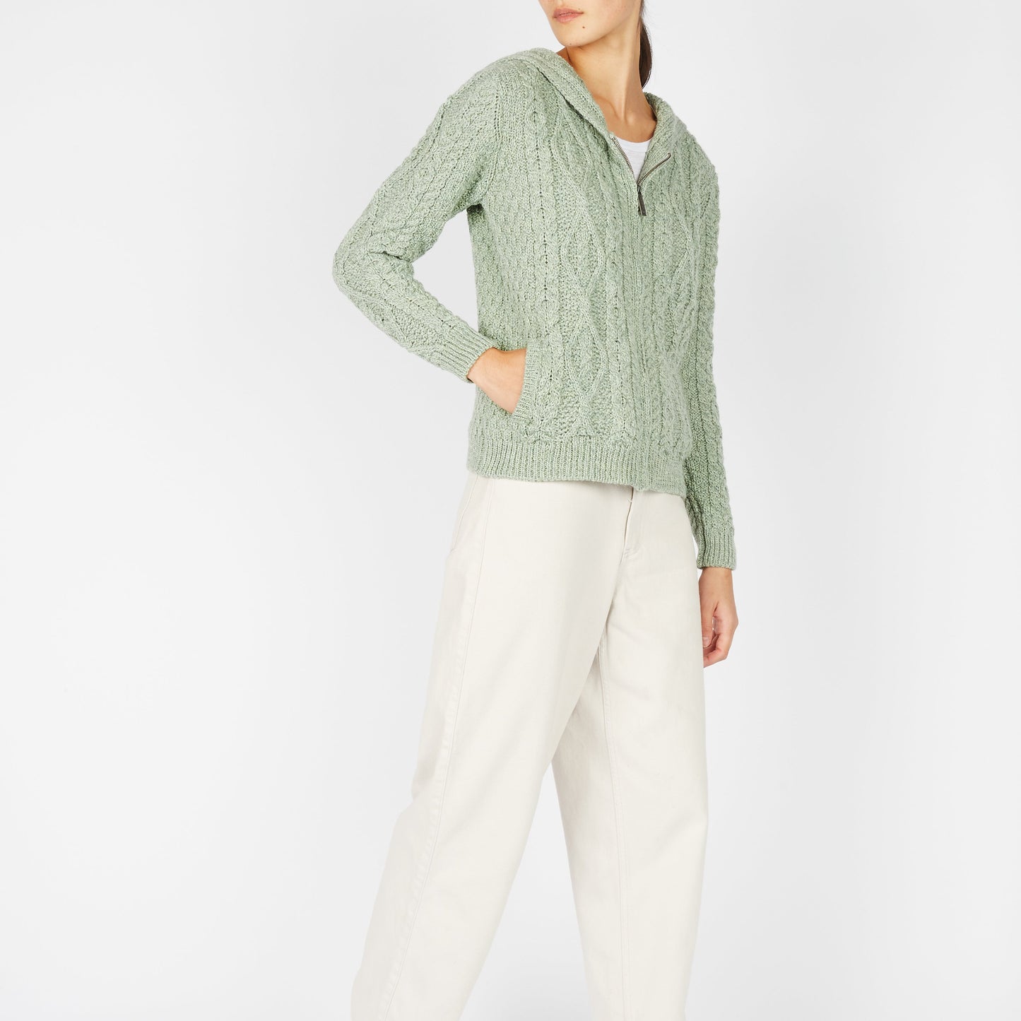 Person wearing a green cable knit sweater and white pants on a light gray background