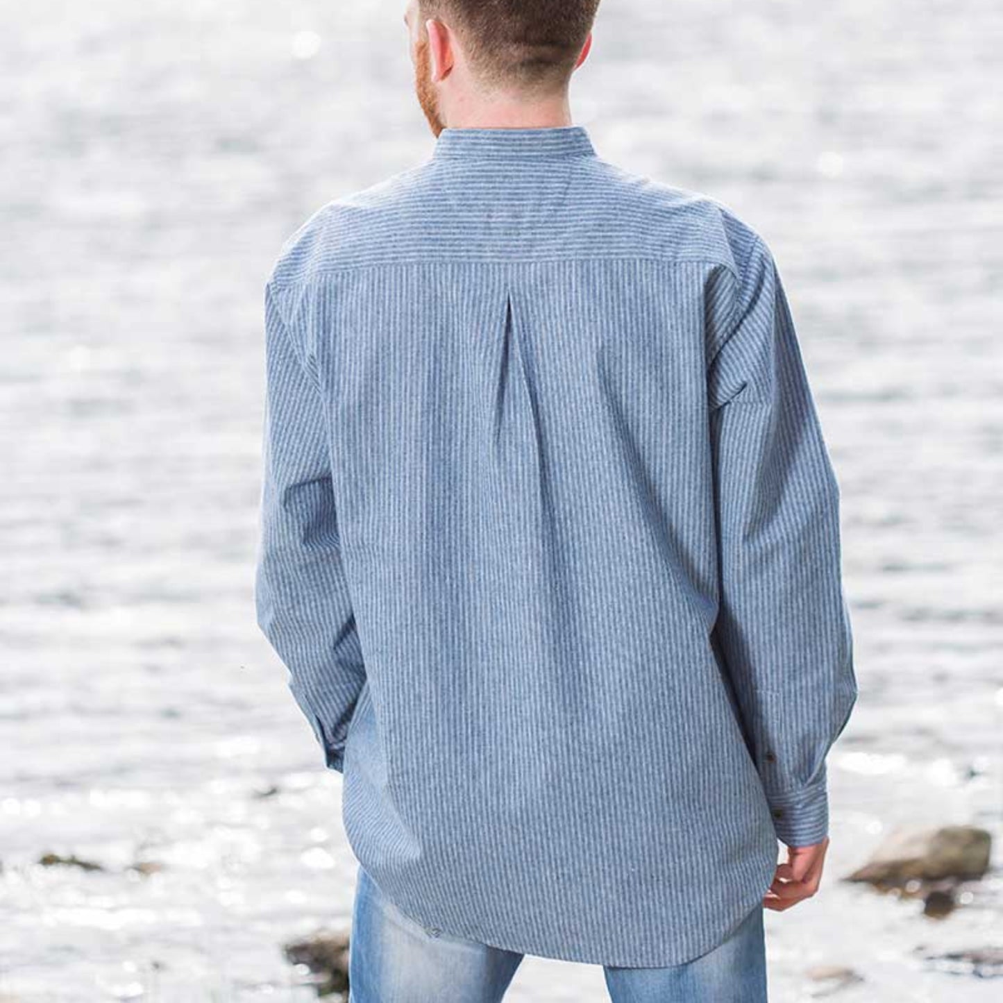 Man standing by a lake wearing a blue checkered shirt with mountains in the background