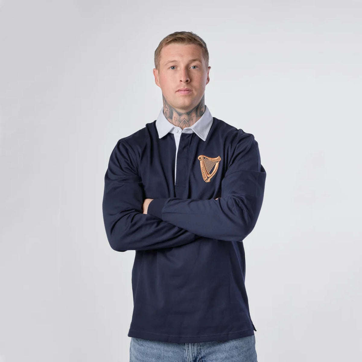 A man wearing a navy blue long-sleeve rugby shirt with a white collar and a gold harp emblem on the chest, standing against a light gray background.