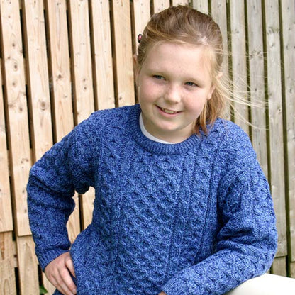 A young girl with light brown hair tied back, wearing a textured blue cable-knit sweater and dark pants, leaning against a white container in front of a wooden fence.