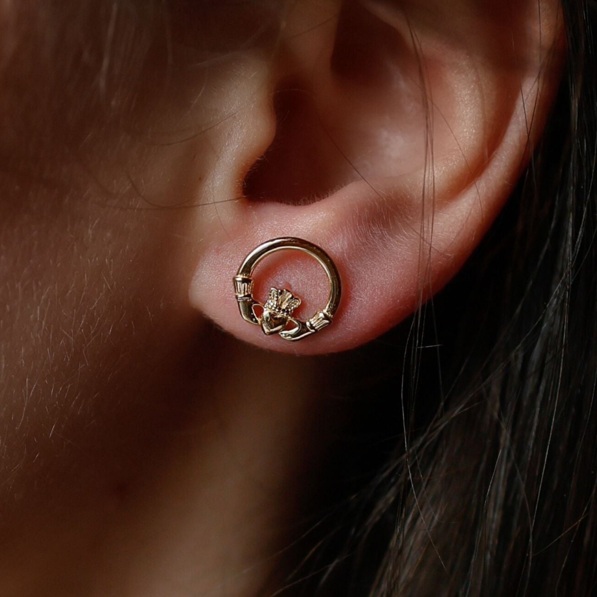 Close-up of a woman's ear with a golden claddagh ring-shaped stud earring.
