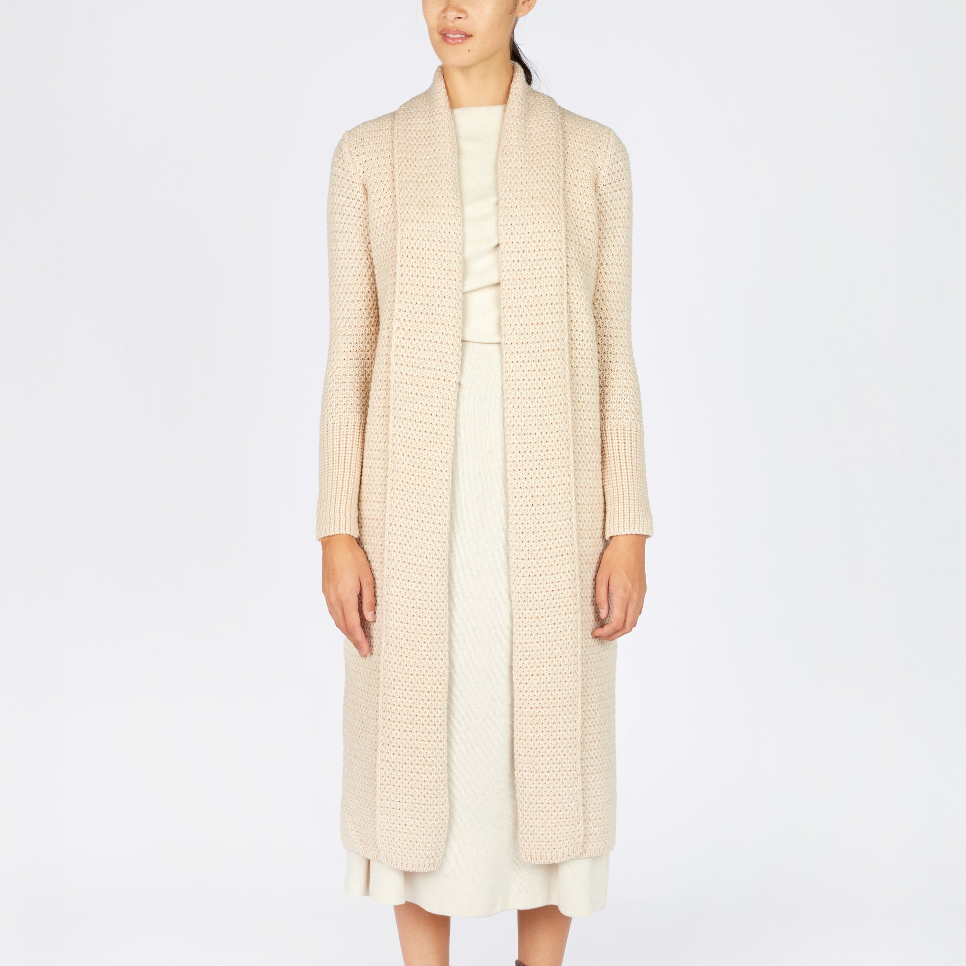 Person wearing a long beige coat over a white dress with brown boots on a white background