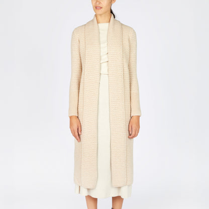 Person wearing a long beige coat over a white dress with brown boots on a white background