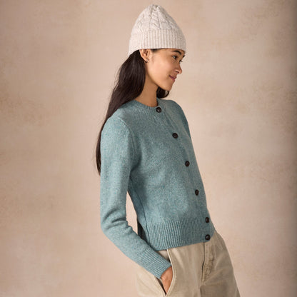 Woman wearing a light blue cardigan with black buttons, beige pants, and a white knit beanie against a beige background.
