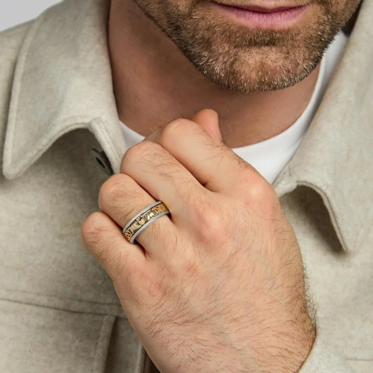 A close-up of a man's hand wearing a two-tone ring with a textured gold inlay, dressed in a light beige jacket over a white t-shirt.