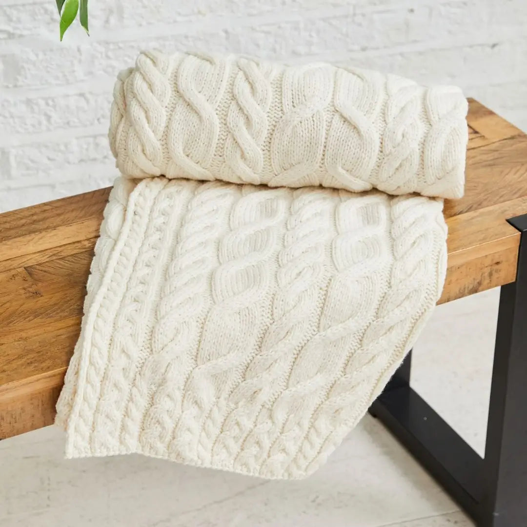 An off-white cable-knit throw blanket draped over a rustic wooden bench against a white brick wall background.
