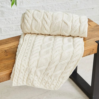 An off-white cable-knit throw blanket draped over a rustic wooden bench against a white brick wall background.