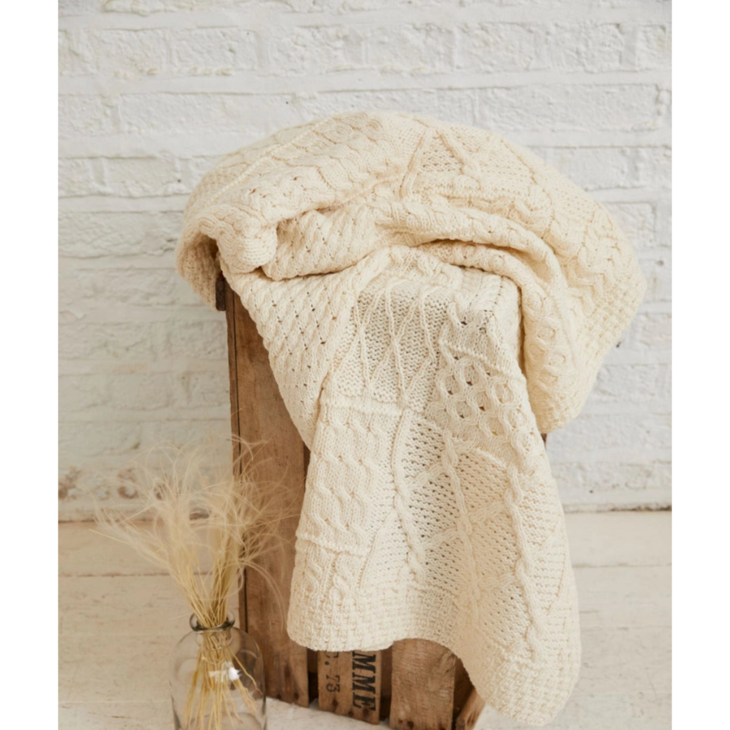 Beige knitted blanket draped over a wooden crate with a small vase of dried grasses on a light wooden floor.