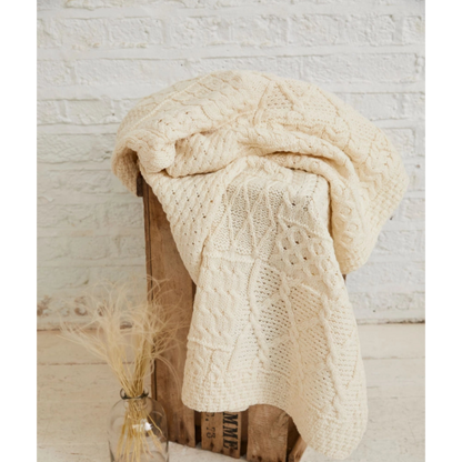 Beige knitted blanket draped over a wooden crate with a small vase of dried grasses on a light wooden floor.