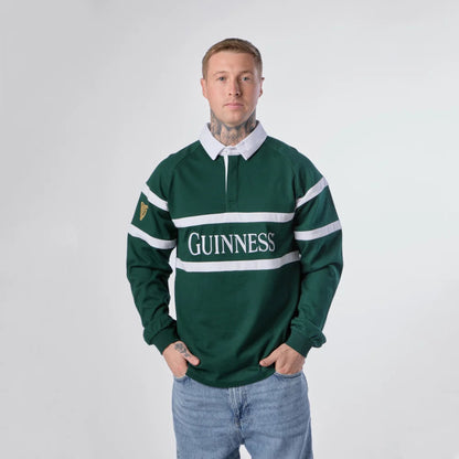 A man wearing a dark green Guinness rugby pullover with white stripes and a white collar, paired with light blue jeans, standing against a light gray studio background.