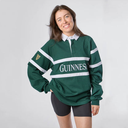 A woman modeling a dark green Guinness rugby shirt with white stripes and a white collar, paired with black bike shorts, against a light gray studio background.