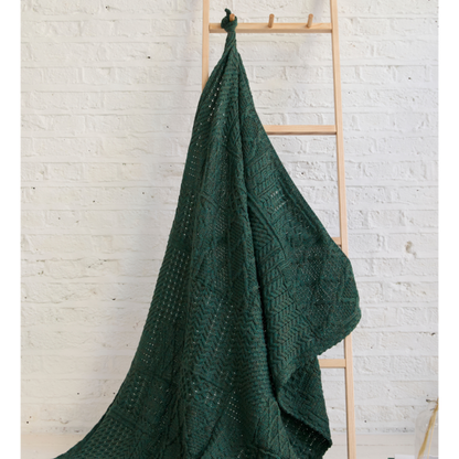 Green knitted blanket draped over a wooden ladder against a white brick wall.
