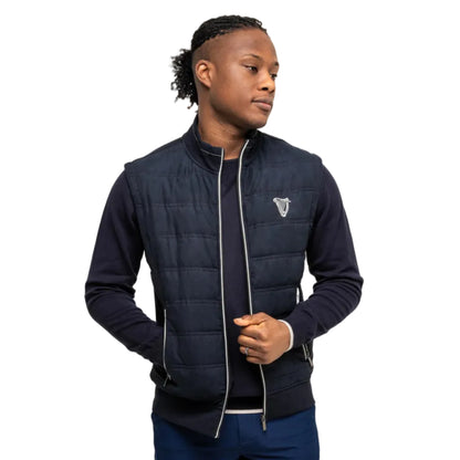 Man wearing a navy-colored quilted vest with a white harp on the front. 