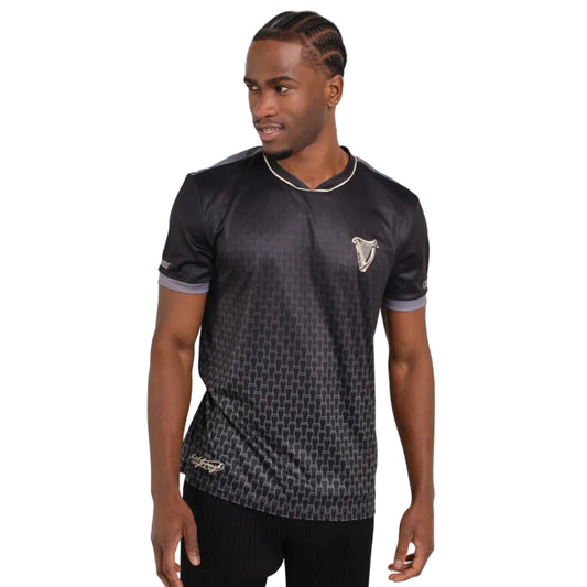 Man wearing a black soccer jersey with a tiny pint glass ombre print and a harp on the left chest.