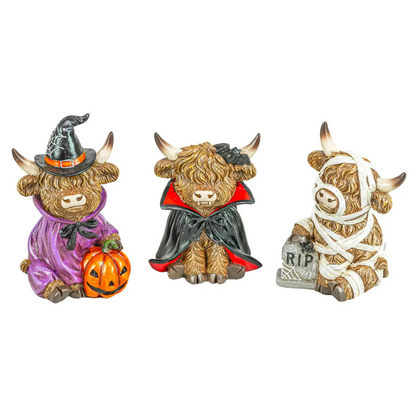 Three Highland cow figurines dressed in Halloween costumes on a plain white background: one in a purple witch outfit with a black hat and holding an orange pumpkin, one in a black and red vampire cape, and one wrapped in white bandages with a small gray RIP grave marker.