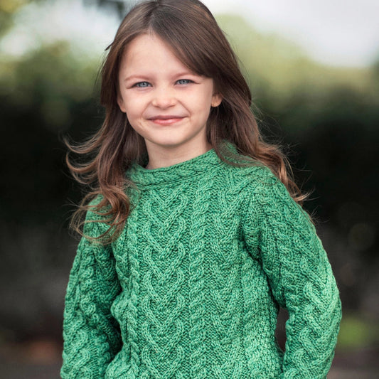 A young girl with long brown hair wearing a green cable-knit sweater and blue jeans, standing outdoors with a blurred natural background.