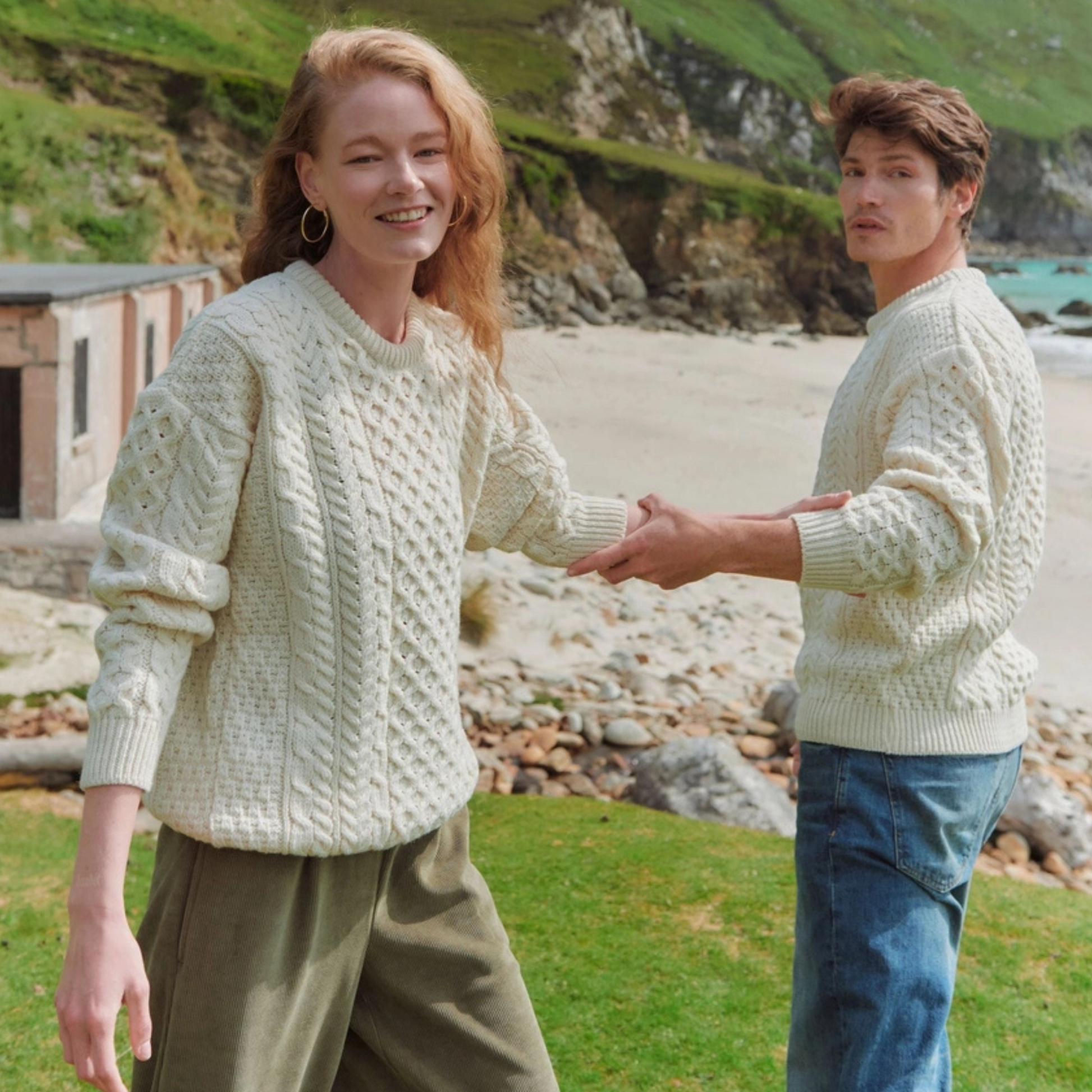 Two people wearing light-colored knitted sweaters on a beach with greenery in the background.