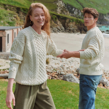 Two people wearing light-colored knitted sweaters on a beach with greenery in the background.
