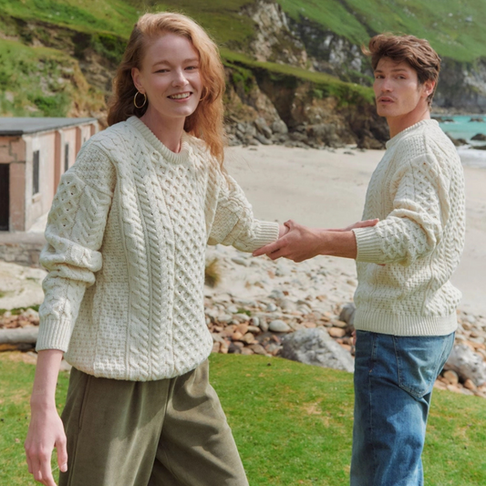 Two people wearing light-colored knitted sweaters on a beach with greenery in the background.