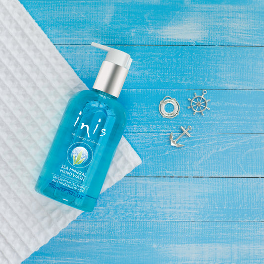 A pump bottle of Inis Sea Mineral Hand Wash on a blue wooden surface, accompanied by a white checkered towel and small decorative nautical items.