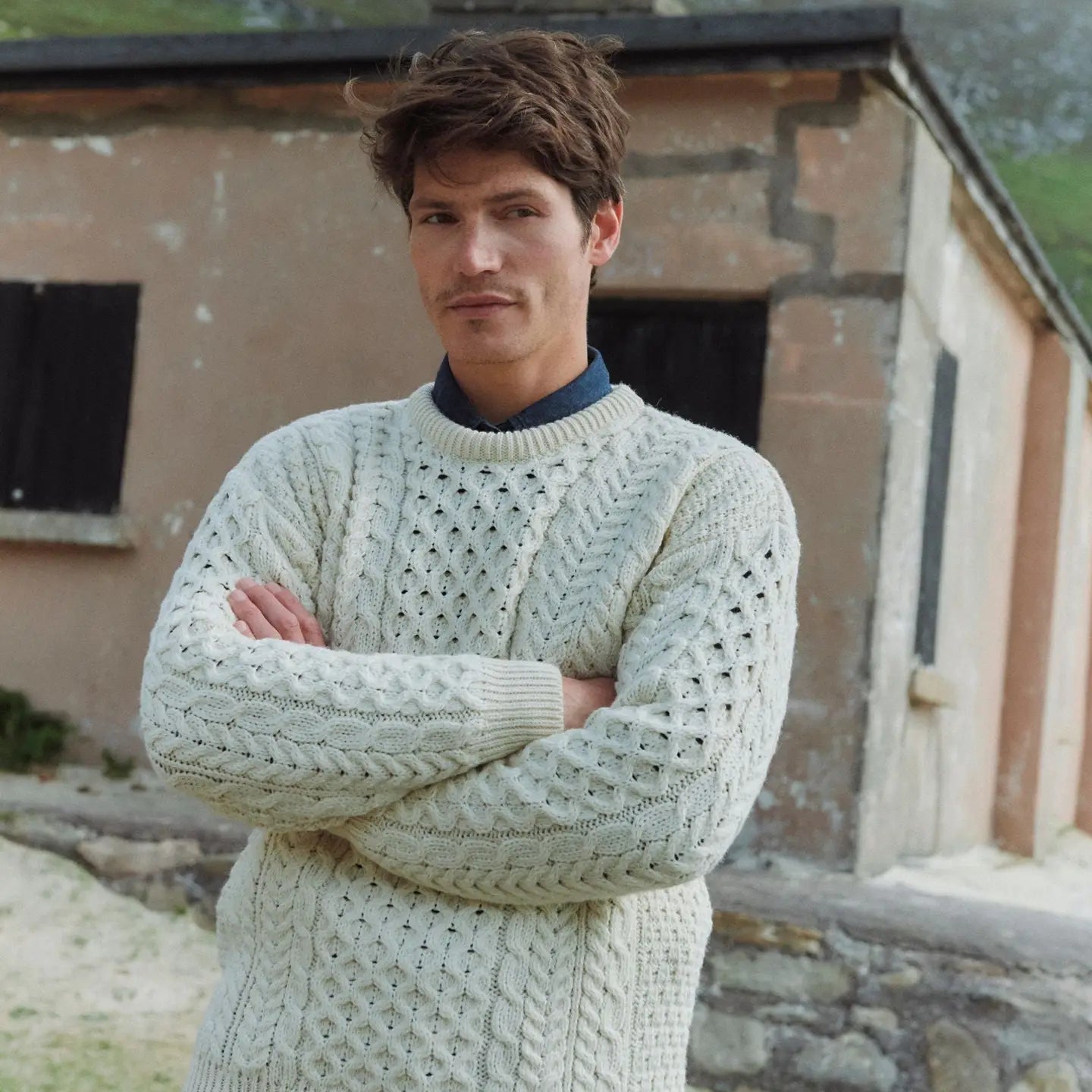 Man wearing a light-colored knitted sweater standing in front of a rustic building with mountains in the background.