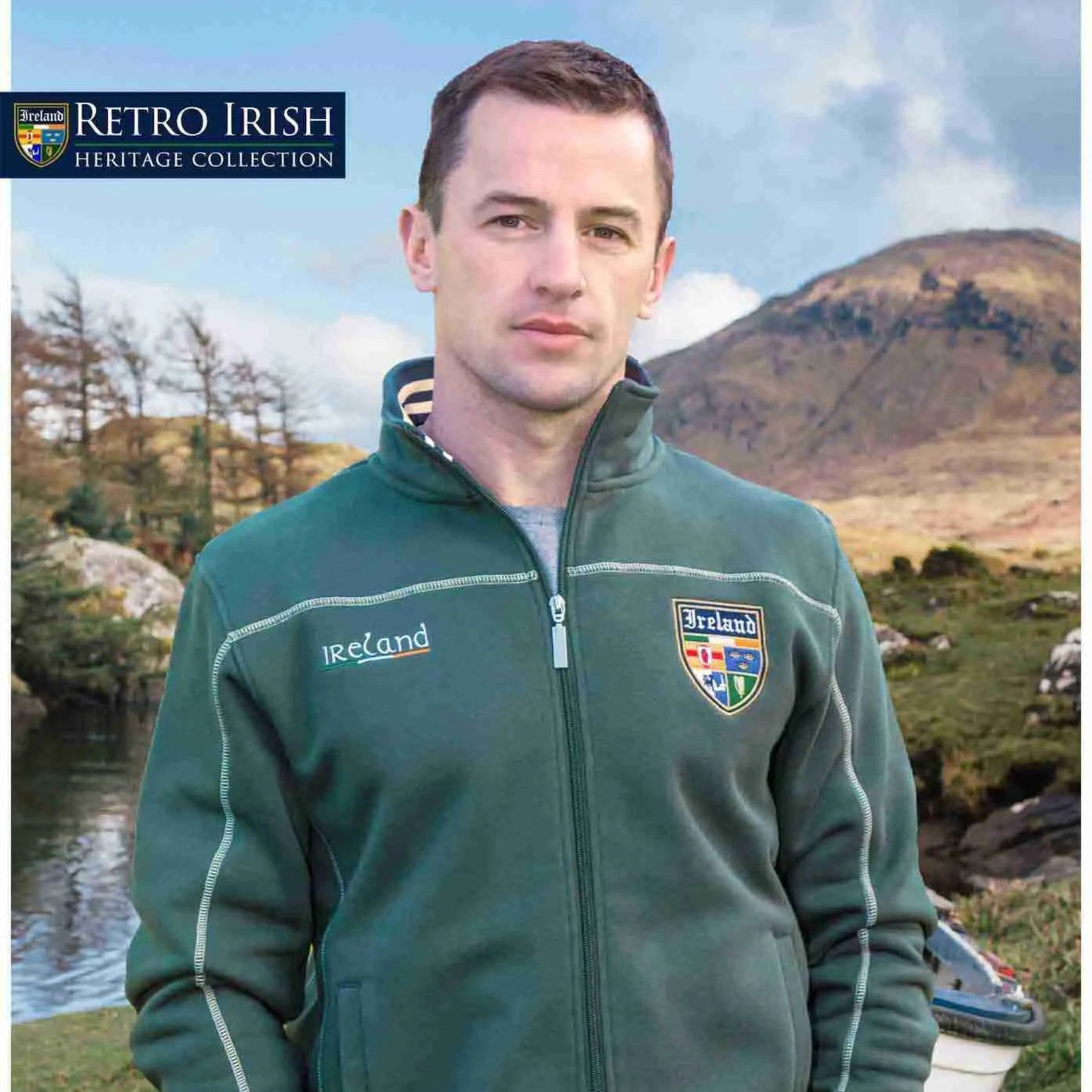Man wearing a green jacket with 'Ireland' branding outdoors near a lake and mountains.