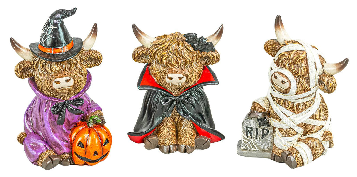 Three Scottish highland cow Halloween figurines on a white background: one in a purple witch outfit holding a pumpkin, one in a black and red vampire cape, and one wrapped in white bandages holding a RIP grave marker.