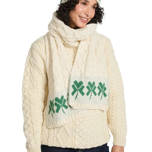 Woman wearing a cream cable knit sweater with green shamrock design and matching scarf on a white background