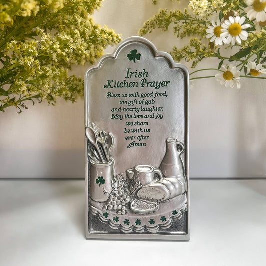 A silver metal plaque with Irish kitchen prayer and decorative elements such as a pot, kettle, and bread.