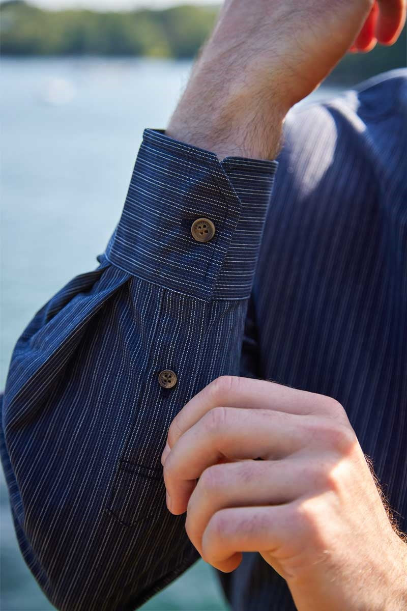 Close-up of a person wearing a blue pinstripe shirt with buttons, outdoors by a body of water.