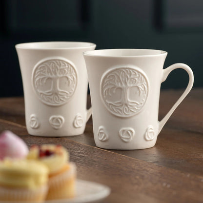 Two white porcelain mugs with tree of life designs on a wooden surface.