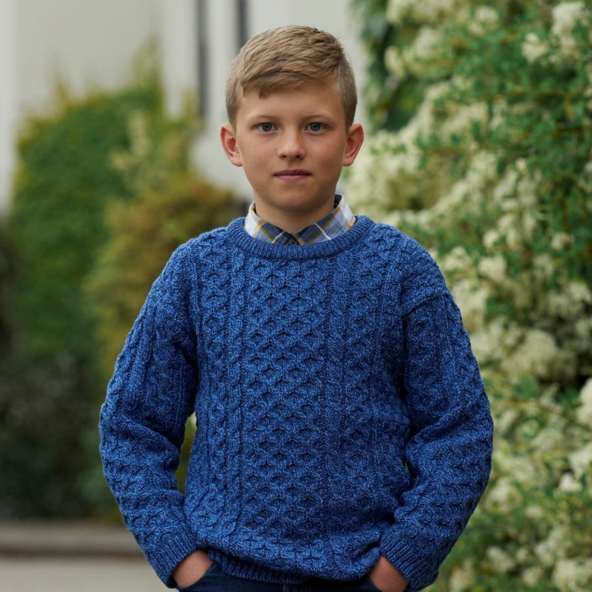 A young boy wearing a textured blue cable-knit sweater over a collared shirt and jeans, standing outdoors with a blurred garden background.