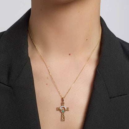 A gold cross pendant necklace with a small green and white stone detail, worn by a model in a black blazer.