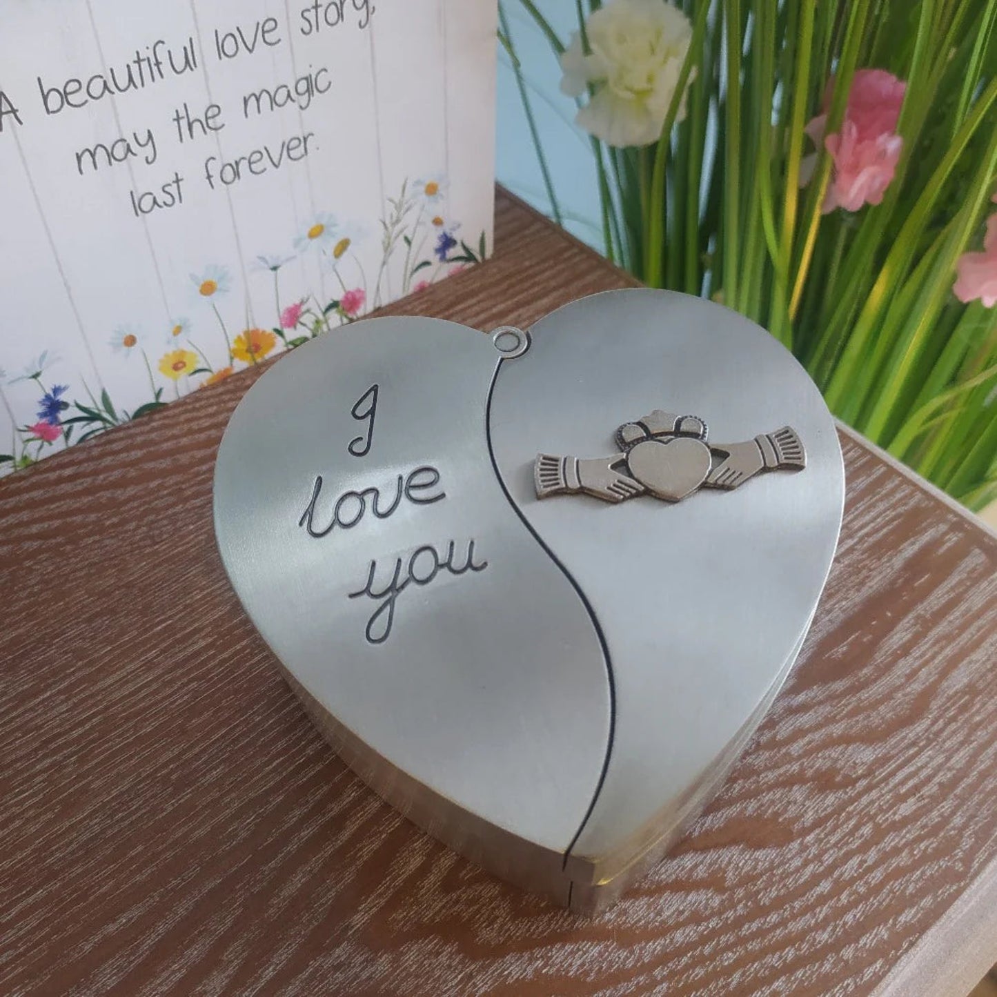 A heart-shaped pewter jewelry box with the Claddagh symbol and the embossed text 'I love you'. The box is placed on a wooden surface with a floral patterned background.