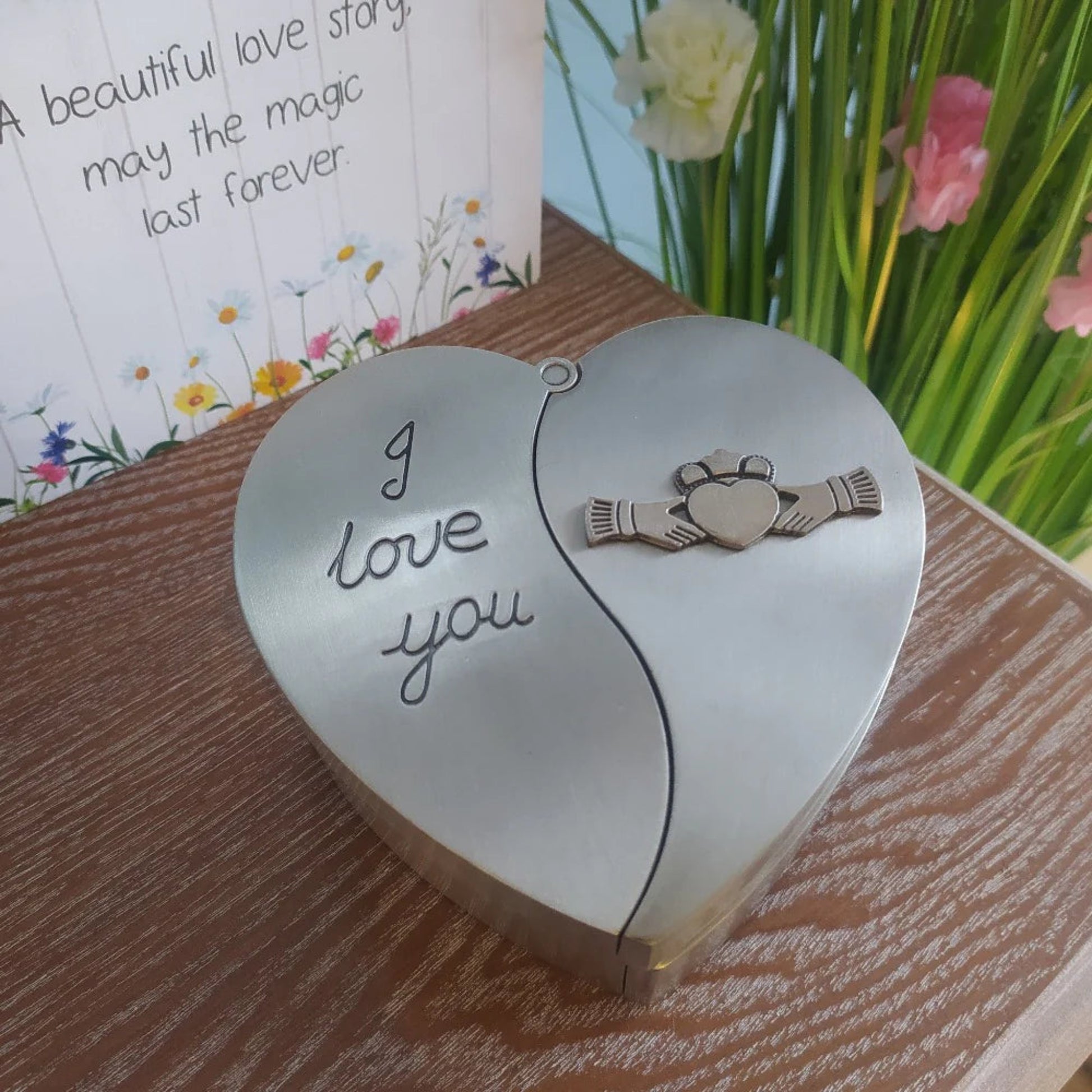 A heart-shaped pewter jewelry box with the Claddagh symbol and the embossed text 'I love you'. The box is placed on a wooden surface with a floral patterned background.