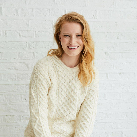 Woman wearing a cream sweater sitting on a stool against a white brick wall.